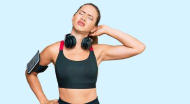 Beautiful blonde woman wearing gym clothes and using headphones suffering of neck ache injury, touching neck with hand, muscular pain 
