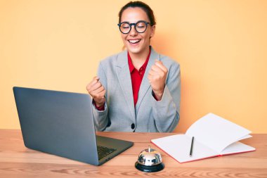 Genç esmer kadın otel resepsiyonunda dizüstü bilgisayar kullanarak çalışıyor. Kolları havada ve gözleri kapalı bir şekilde başarıyı kutluyor ve şaşırıyor. 