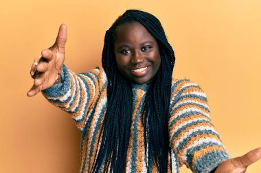 Saçları örgülü genç siyahi bir kadın sıradan bir kazak giyiyor ve kameraya sarılıp gülümsüyor. Mutluluğu kucaklayan neşeli bir ifade.. 