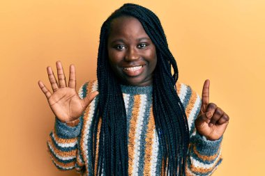Saçları örgülü genç siyahi bir kadın günlük kışlık süveter giyiyor ve altı numaralı parmaklarıyla işaret ederken kendinden emin ve mutlu bir şekilde gülümsüyor.. 