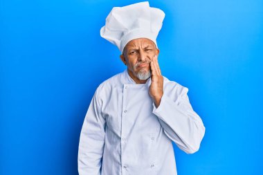 Orta yaşlı, gri saçlı, profesyonel aşçı üniforması giyen ve diş ağrısı ya da diş hastalığından dolayı ağızlarına acı dolu bir ifadeyle dokunan şapkalı bir adam. dişçi 