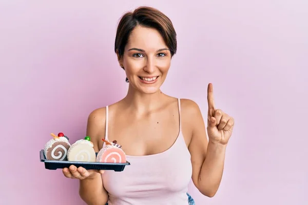 若いですブルネット女性ともに短い髪を保持ケーキお菓子笑顔でアイデアや質問を指差す指で幸せな顔 ナンバーワン — ストック写真