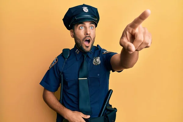 Handsome hispanic man wearing police uniform pointing with finger surprised ahead, open mouth amazed expression, something on the front
