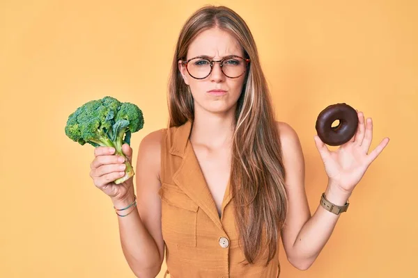 Young Blonde Girl Holding Broccoli Chocolate Donut Skeptic Nervous Frowning — Stock Photo, Image