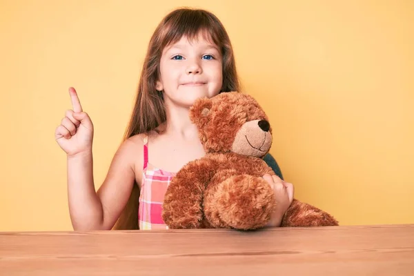小さなCaucasian子供の女の子とともに長いです髪座っているテーブルの上にテディベア笑顔幸せなポインティングで手と指で側面 — ストック写真