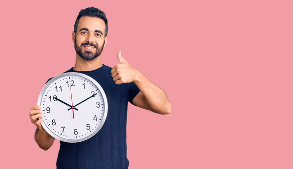 Joven Hombre Hispano Sosteniendo Gran Reloj Sonriendo Feliz Positivo Pulgar — Foto de Stock