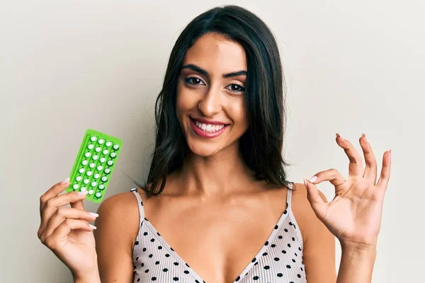 Beautiful hispanic woman holding birth control pills doing ok sign with fingers, smiling friendly gesturing excellent symbol