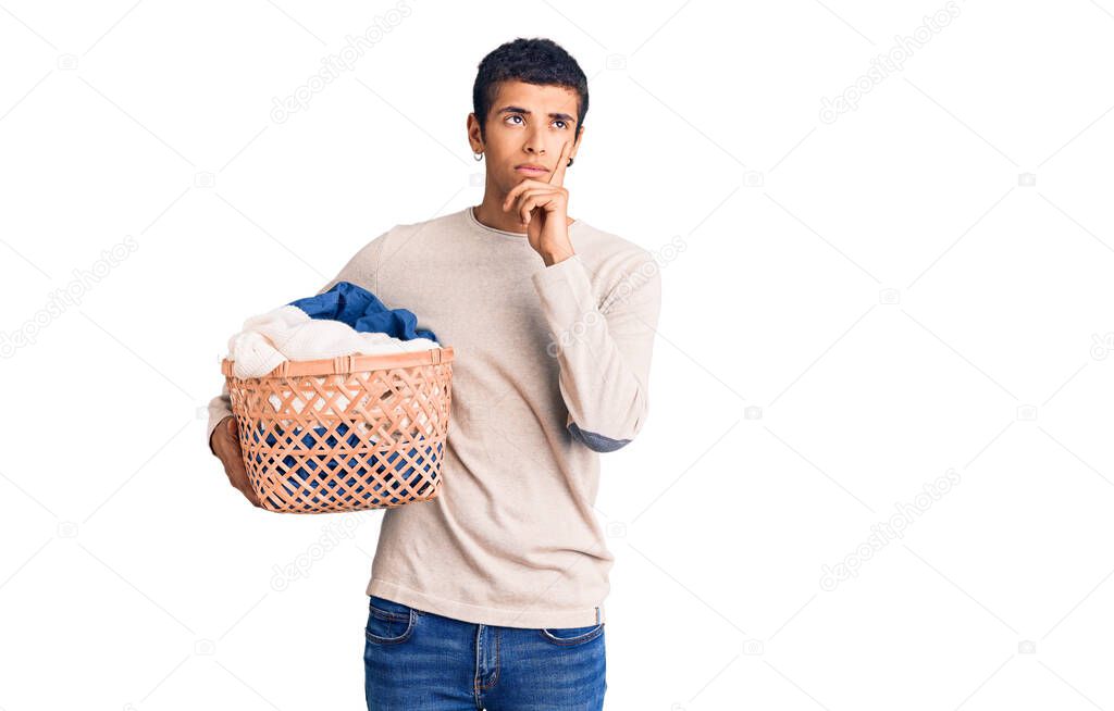 Young african amercian man holding laundry basket serious face thinking about question with hand on chin, thoughtful about confusing idea 
