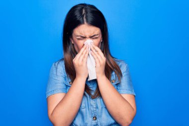 Genç ve güzel bir kadın gribe yakalandı. İzole edilmiş mavi arka planda duran sümük dolu burun üzerine kağıt mendil kullanarak.