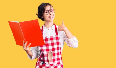 Kısa saçlı, profesyonel fırıncı önlüğü takan, yemek tarifi kitabı okuyan mutlu ve pozitif gülümseyen, başparmağını kaldıran ve onay veren güzel bir kadın. 