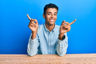 Genç, yakışıklı, Afro-Amerikalı, gündelik giysiler giymiş masada oturmuş, kendine güvenen, parmaklarıyla farklı yönleri gösteren bir şekilde gülümsüyor. reklam için kopyalama alanı 