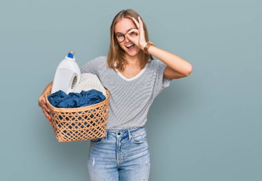 Çamaşır sepeti ve deterjan şişesi tutan güzel sarışın kadın gülümsüyor. 