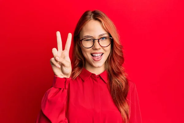 年轻美丽的红头发女人穿着休闲装 戴着眼镜 穿着红色背景的衣服 高兴地微笑着 脸上闪烁着胜利的标志 第二点 — 图库照片