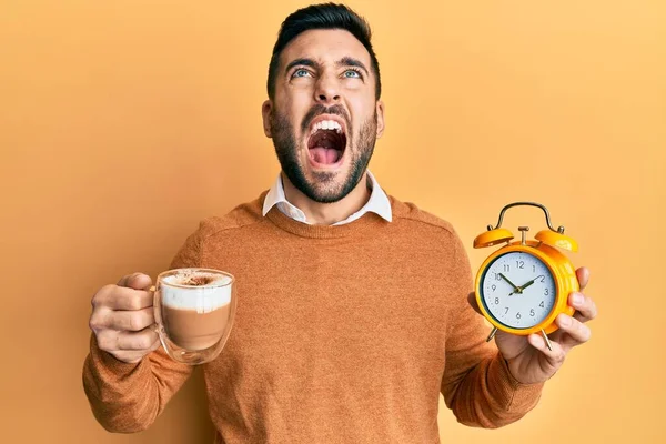 Young hispanic man holding coffee and alarm clock angry and mad screaming frustrated and furious, shouting with anger looking up.