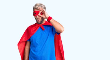 Süper kahraman gibi giyinmiş genç sarışın adam şaşırmış yüz ifadesiyle şaşırmış, parmaklarının arasından bakan bir hareket yapıyor. İnançsız ifade. 