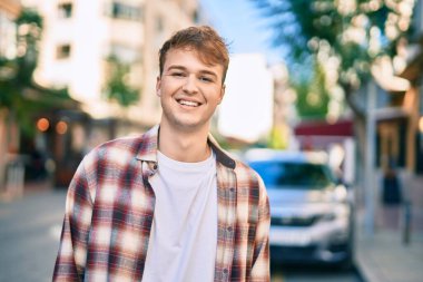 Genç beyaz adam gülümseyerek şehirde mutlu mesut duruyor..