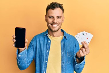 Poker kartları ve akıllı telefon tutan genç İspanyol adam yüzünde mutlu ve havalı bir gülümsemeyle boş ekranı gösteriyor. diş gösterme. 