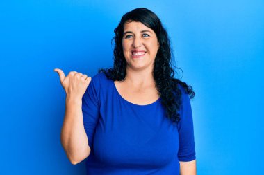 Artı mavi bir gömlek giyen esmer kadın mutlu yüzüyle gülümseyen ve baş parmağıyla yan tarafı gösteren.. 