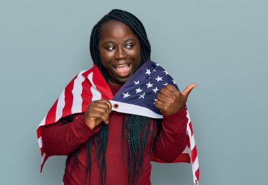 Birleşik Devletler bayrağını tutan siyah örgülü genç bir kadın başparmağını yana kaldırmış, ağzı açık gülümsüyor. 