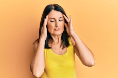 Orta yaşlı esmer kadın sarı arka planda gündelik kıyafetler giyiyor. Başı ağrıyor çünkü stres altında. migren ağrısı. 