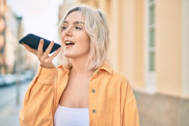 Genç sarışın kız gülümseyerek şehirde akıllı telefon kullanarak sesli mesaj gönderiyor..