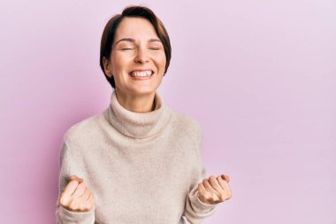 Kısa saçlı esmer kadın, günlük kışlık süveter giyiyor. Çok mutlu ve heyecanlı. Kolları havada, gülümseyerek ve başarı için çığlık atarak kazanan jest yapıyor. kutlama konsepti. 