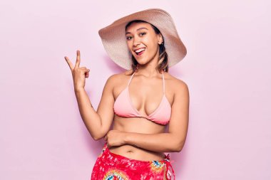 Bikini giyen, şapkalı, gülümseyen ve kameraya göz kırpan genç bir kadın zafer işareti yapıyor. İki numara.. 