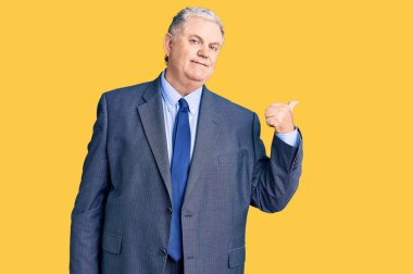 İş ceketi giyen, gülümseyen, gülümseyen ve baş parmağıyla yüzünü gösteren yaşlı, gri saçlı bir adam.. 