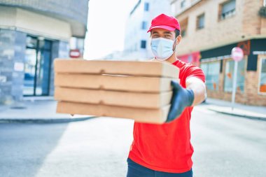 Üniforma giyen ve koronavirüs koruma maskesi takan genç teslimatçı. Kasaba caddesinde pizza kutularını teslim ediyorum..