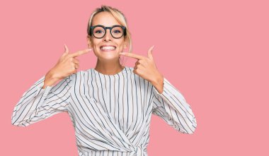 İş gömleği ve gözlük takan güzel sarışın kadın neşeli bir şekilde gülümsüyor ve parmakları ve ağızlarıyla işaret ediyor. diş sağlığı kavramı. 