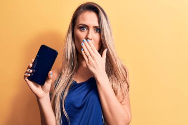 Sarışın, güzel bir kadın elinde akıllı telefonuyla sarı arka planda ağzı elle kaplayan, şok olmuş ve hata yapmaktan korkan bir ekran gösteriyor. Şaşırtıcı ifade.