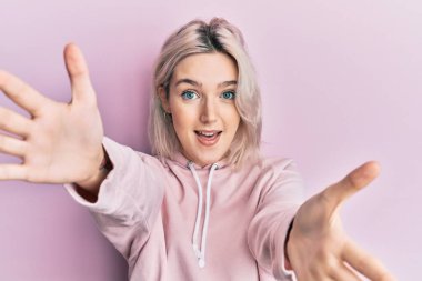 Sıradan kıyafetler giyen genç sarışın kız kameraya bakıp sarılmak için kollarını açmış gülümsüyor. Mutluluğu kucaklayan neşeli bir ifade.. 