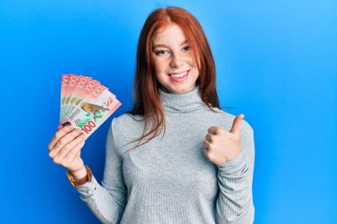 Young red head girl holding 100 new zealand dollars banknote smiling happy and positive, thumb up doing excellent and approval sign 