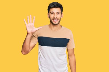 Gündelik kıyafetler giyen genç yakışıklı adam beş numarayı gösterip işaret ederken kendinden emin ve mutlu bir şekilde gülümsüyor.. 