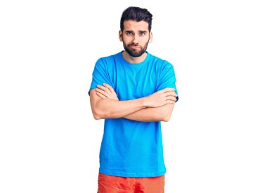Sakallı, sıradan tişörtlü, kuşkucu ve gergin, yüzünde çapraz kollu tasvip etmeyen bir ifade olan genç yakışıklı bir adam. negatif kişi. 