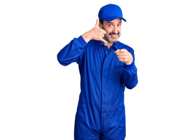 Middle age handsome man wearing mechanic uniform smiling doing talking on the telephone gesture and pointing to you. call me.  clipart