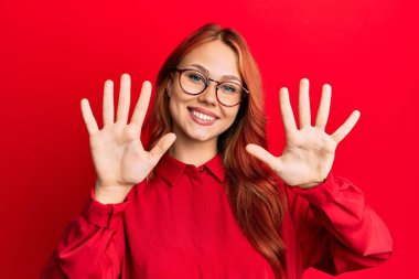 Kırmızı arka planda günlük kıyafetler ve gözlük takan genç, güzel bir kızıl kadın on numarayı gösterirken kendinden emin ve mutlu bir şekilde gülümsüyor.. 