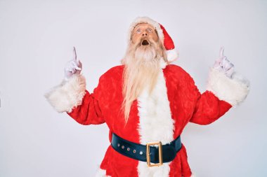 Gri saçlı, uzun sakallı, Noel Baba kostümü giyen yaşlı bir adam şaşırmış ve şaşırmış bir şekilde yukarı bakıyor ve parmaklarıyla işaret ediyor ve kollarını kaldırıyor.. 