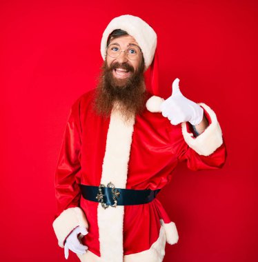 Uzun sakallı yakışıklı kızıl saçlı Noel Baba kostümü giyen mutlu ve pozitif gülümseyen mükemmel ve onay işareti yapan genç adam. 
