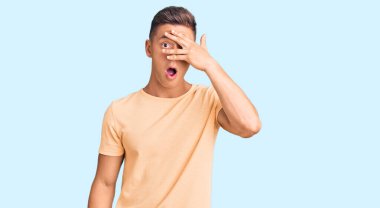 Gündelik kıyafetler giyen genç yakışıklı adam şok içinde yüzünü ve gözlerini kapayarak utangaç ifadesiyle parmaklarının arasından bakıyor.. 