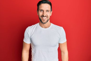 Yüzünde mutlu ve havalı bir gülümseme olan günlük beyaz tişört giyen genç yakışıklı bir adam. şanslı kişi. 