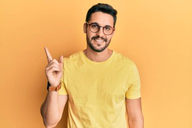 Gündelik kıyafetler ve gözlük takan, yüzünde kocaman bir gülümseme olan, eli ve parmağı kameraya bakan genç İspanyol bir adam.. 