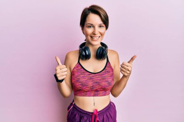 Kısa saçlı, spor giyimli ve kulaklık takan genç esmer bir kadın. Başparmağıyla gülümseyen ve mutlu bir şekilde olumlu jest yapıyor. Neşeli ifade ve kazanan jest. 