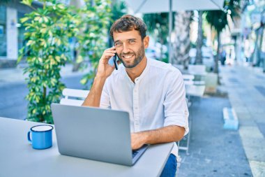 Güneşli bir günde günlük beyaz gömlek giyen sakallı yakışıklı adam laptopla çalışıyor ve kafeteryada telefonla konuşuyor.