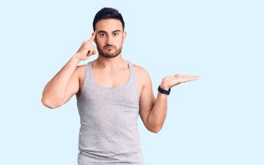 Mayo giyen, kolsuz tişört giyen, kafası karışmış ve avuç içi açık, fotokopi alanı gösteren ve alnını işaret eden genç yakışıklı bir adam. Bunu bir düşün.. 