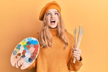 Güzel beyaz bir kız elinde boya fırçası ve ressam paleti ile kızgın ve kızgın çığlıklar atıyor sinirlenmiş ve öfkeli, öfkeyle bağırıyor.. 