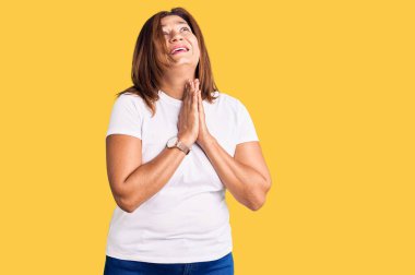 Orta yaşlı, beyaz bir tişört giyen Latin bir kadın elleri bağlı yalvarıyor ve dua ediyor. Yüzünde umut ifadesi var. Çok duygusal ve endişeli. Yalvarıyor. 