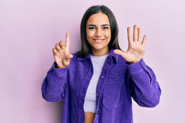 Genç esmer kadın gündelik giysiler giyer ve yedi numaralı parmaklarıyla işaret ederken kendine güvenen ve mutlu bir şekilde gülümser.. 
