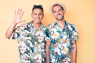 Genç eşcinsel çift, yaz şapkası ve Hawaii gömleği takıyor. Beş numaralı parmaklarıyla işaret ediyor ve gülümsüyor. Kendinden emin ve mutlu.. 
