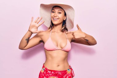 Bikini giyen ve şapka takan genç bir kadın yedinci parmaklarını gösterip işaret ederken kendinden emin ve mutlu bir şekilde gülümsüyor.. 